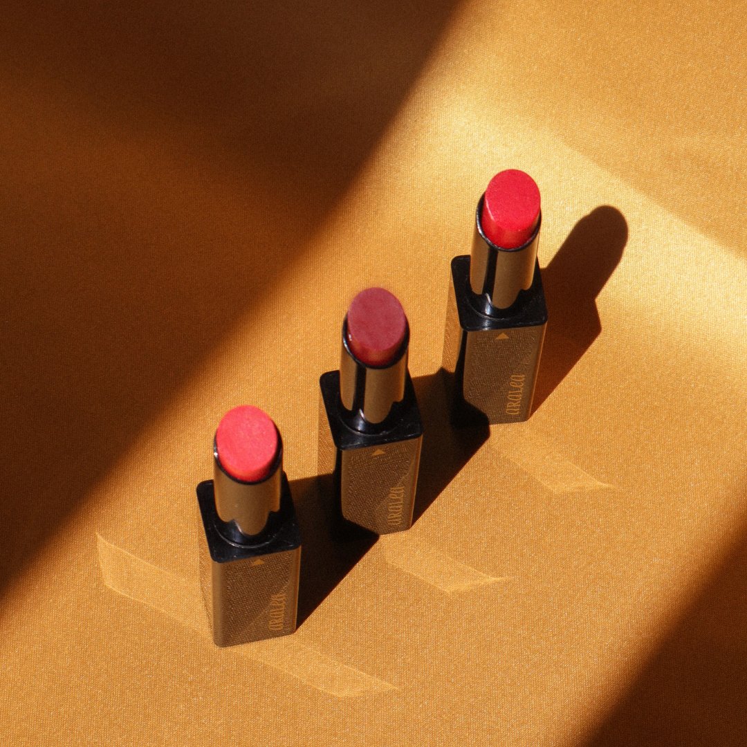 Aerial photo of three SUN KISS TINTS vitamin D lip tints standing upright in a row against deep gold silk fabric in a beam of sunlight.