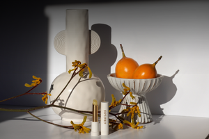Two SUN KISS BALM vitamin D lip balms stand upright on white posterboard in front of a cream ceramic vase, a ceramic pedestal dish with two granadillas, and a stem of yellow flowers.