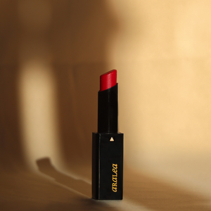SUN KISS TINT RUBY vitamin D infused lip tint stands upright on light gold silk fabric in a beam of sunlight.