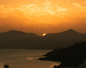 A sun sets behind a mountain range with curving coastline and ocean. The sun is partially obscured by the mountain range and the sky is shades of deep orange and brighter yellow.