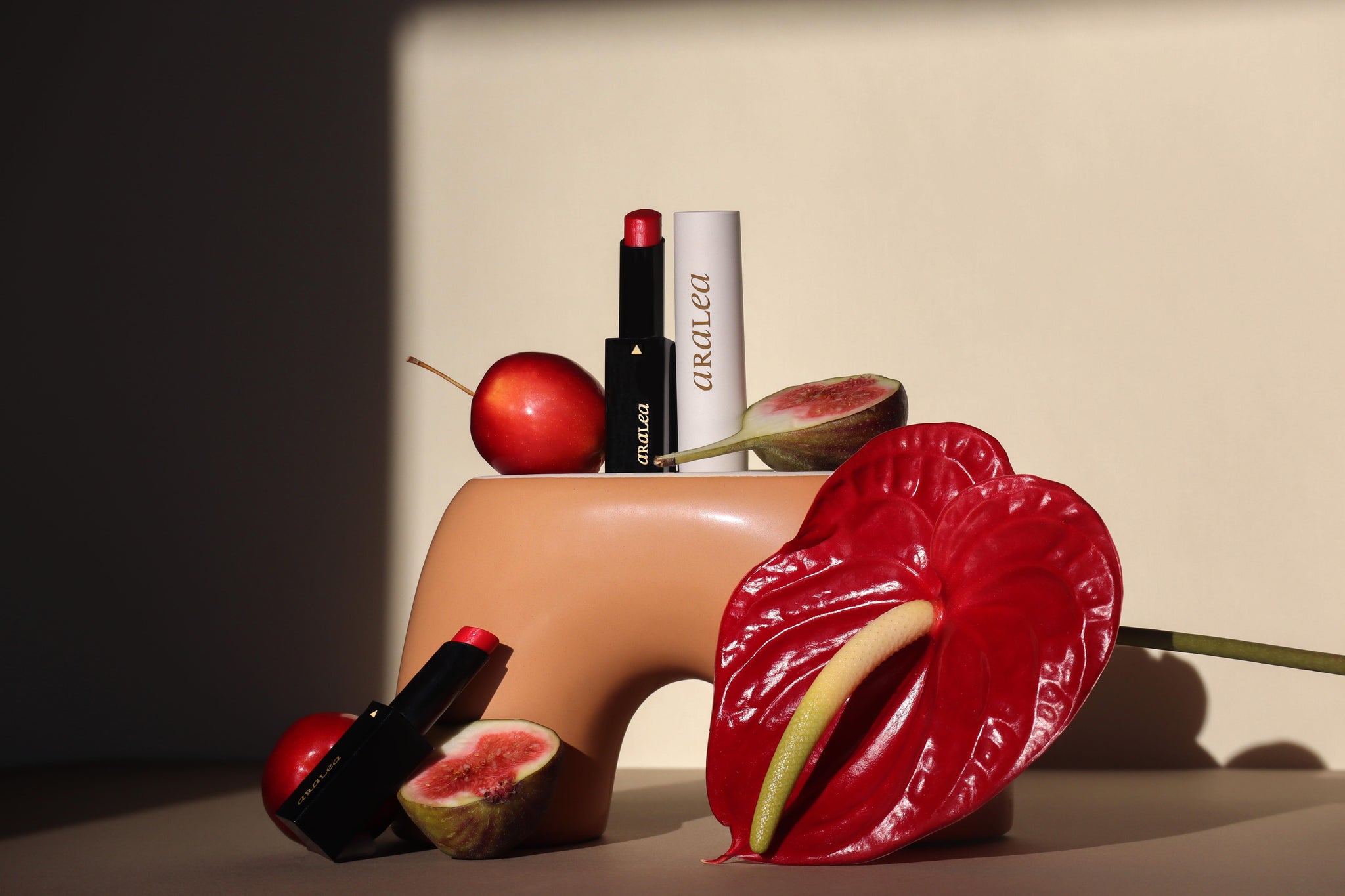 SUN KISS TINTS vitamin D infused lip care are positioned around a terra-cotta coloured ceramic vase nestled against a red anthurium bloom, fresh figs, and a crab apple. In direct sunlight against a beige poster board background.