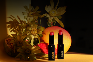 Two SUN KISS TINT vitamin D lip tints stand upright in front of a mango and stems of leucadendron on deep yellow posterboard lit by direct sunlight and intense shadow.