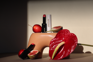 SUN KISS TINTS vitamin D infused lip care are positioned around a terra-cotta coloured ceramic vase nestled against a red anthurium bloom, fresh figs, and a crab apple. In direct sunlight against a beige poster board background.