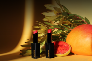 Two SUN KISS TINTS vitamin D lip care stand upright nestled against a mango and pitaya and stems of leucadendron. Positioned on deep yellow-gold posterboard in direct sunlight with intense shadow.