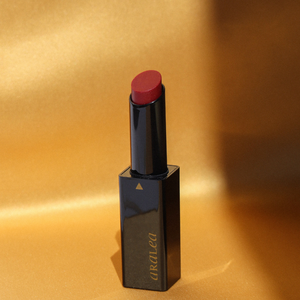 SUN KISS TINT DARK DAHLIA vitamin D infused lip tint standing upright against a background of gold silk standing upright in a beam of sunlight.
