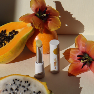 SUN KISS BALM vitamin D infused lip balm stands upright in front of a background of orange tulips, a yellow pitaya fruit, and a passionfruit.
