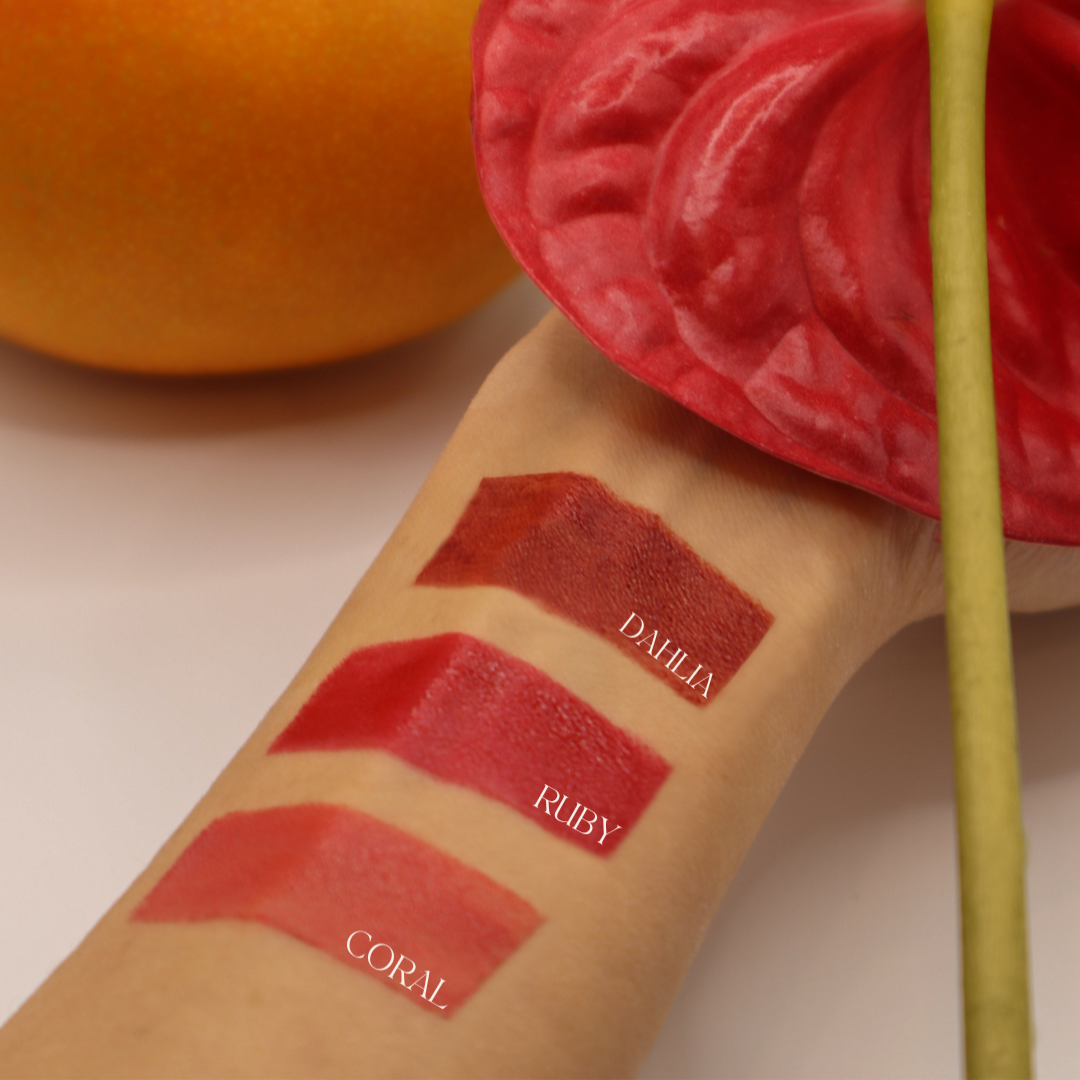 An arm against a plain white background with three SUN KISS TINT swatches swiped across arm surrounded by an orange mango and a red anthurium.