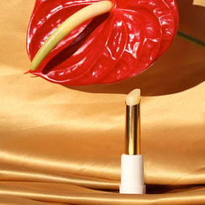 SUN KISS BALM vitamin D infused lip balm stands upright on gold silk fabric in sunlight with a red anthurium bloom